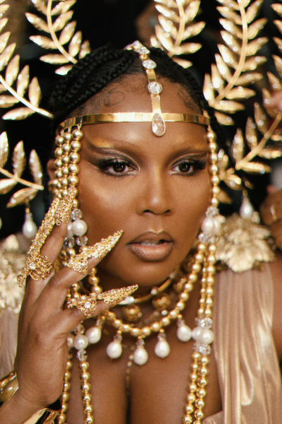 behind the scenes shot of lizzo wearing a golden hairpiece and gold smokey eye with gold stiletto nail jewelry
